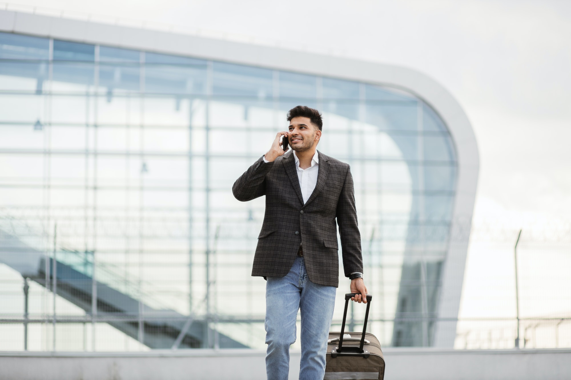 Indian business man walking outside to station or airport talking on the phone and carrying suitcase